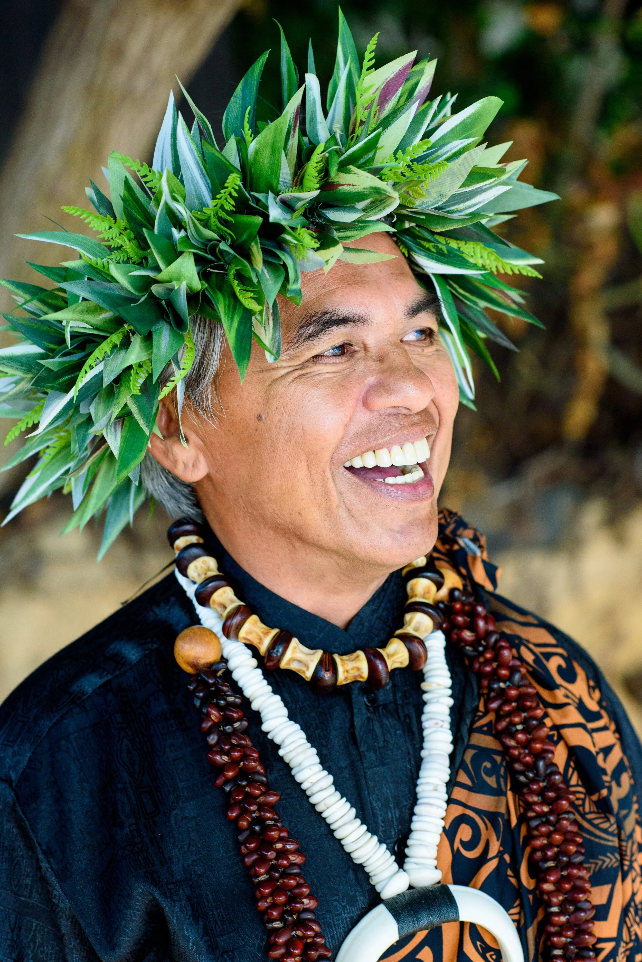 A smiling man wearing traditional Polynesian tribal attire, including a leafy head wreath, multiple beaded necklaces, and a patterned shirt.