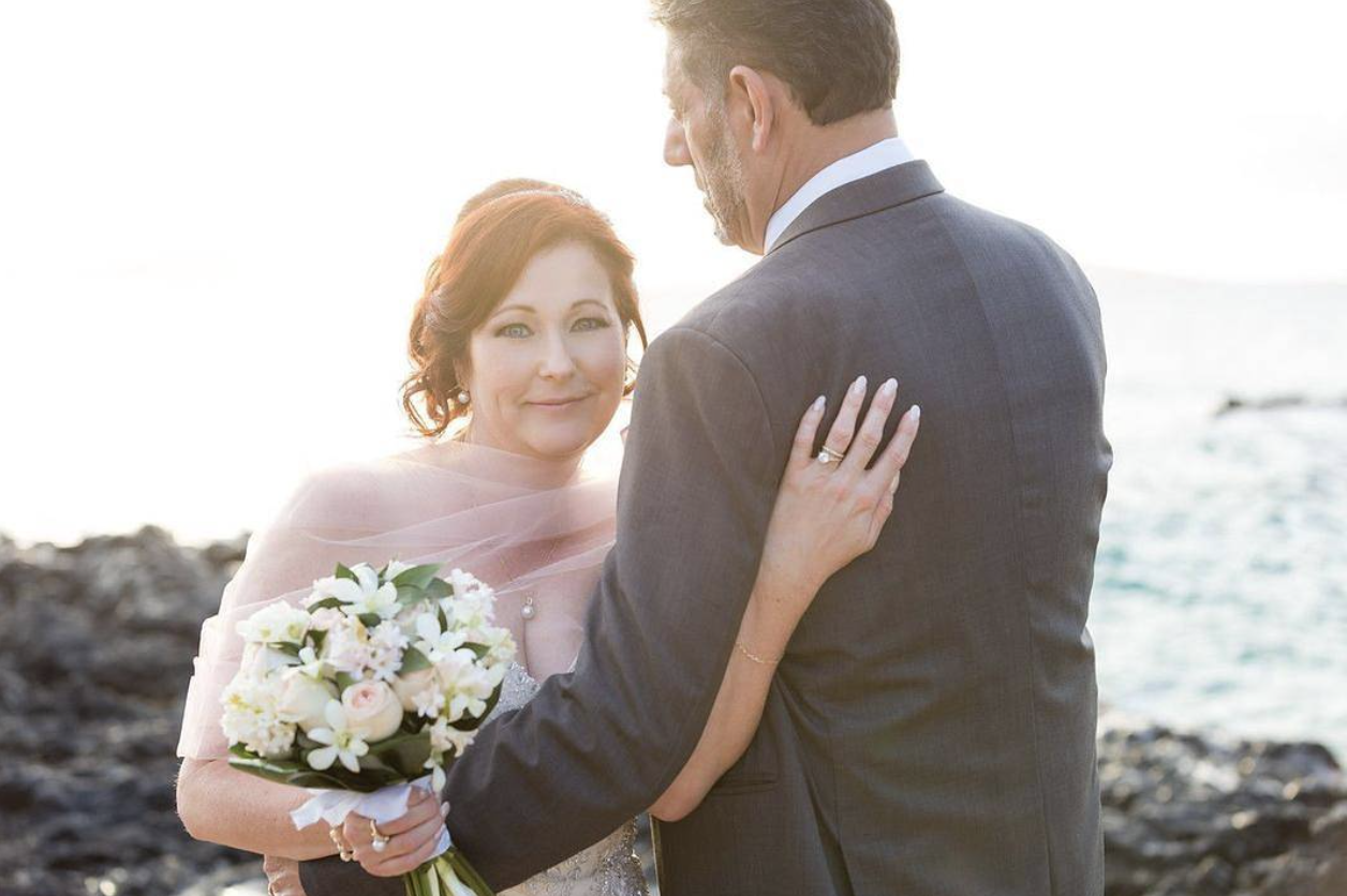 A couple on the beach kissing, with the woman holding a bouquet of flowers and the man in light-colored pants and a white shirt.