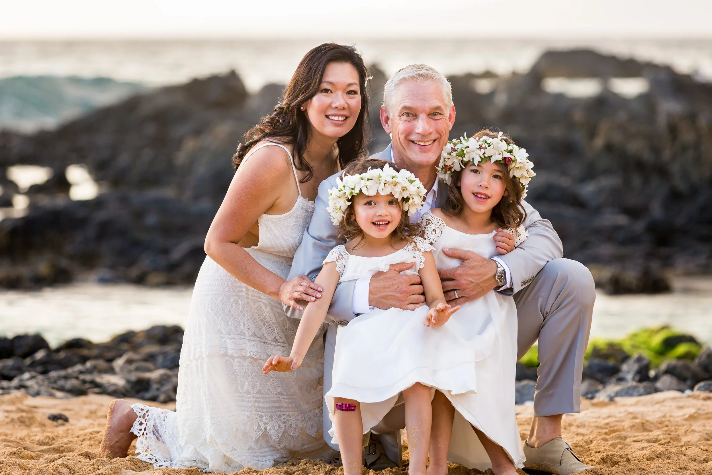 A family celebrating a wedding on the beach. The parents and two young girls are dressed in white, with floral crowns on the girls' heads. They are kneeling and sitting on the sand with rocks and the ocean in the background, smiling at the camera.