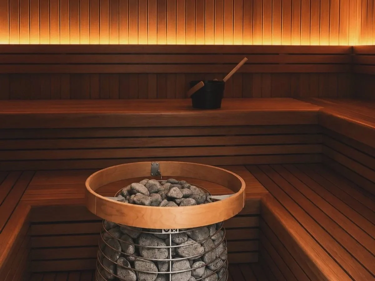 Interior of a wooden sauna with a bucket of water and a ladle on a bench, and stones in a metal basket for heating.