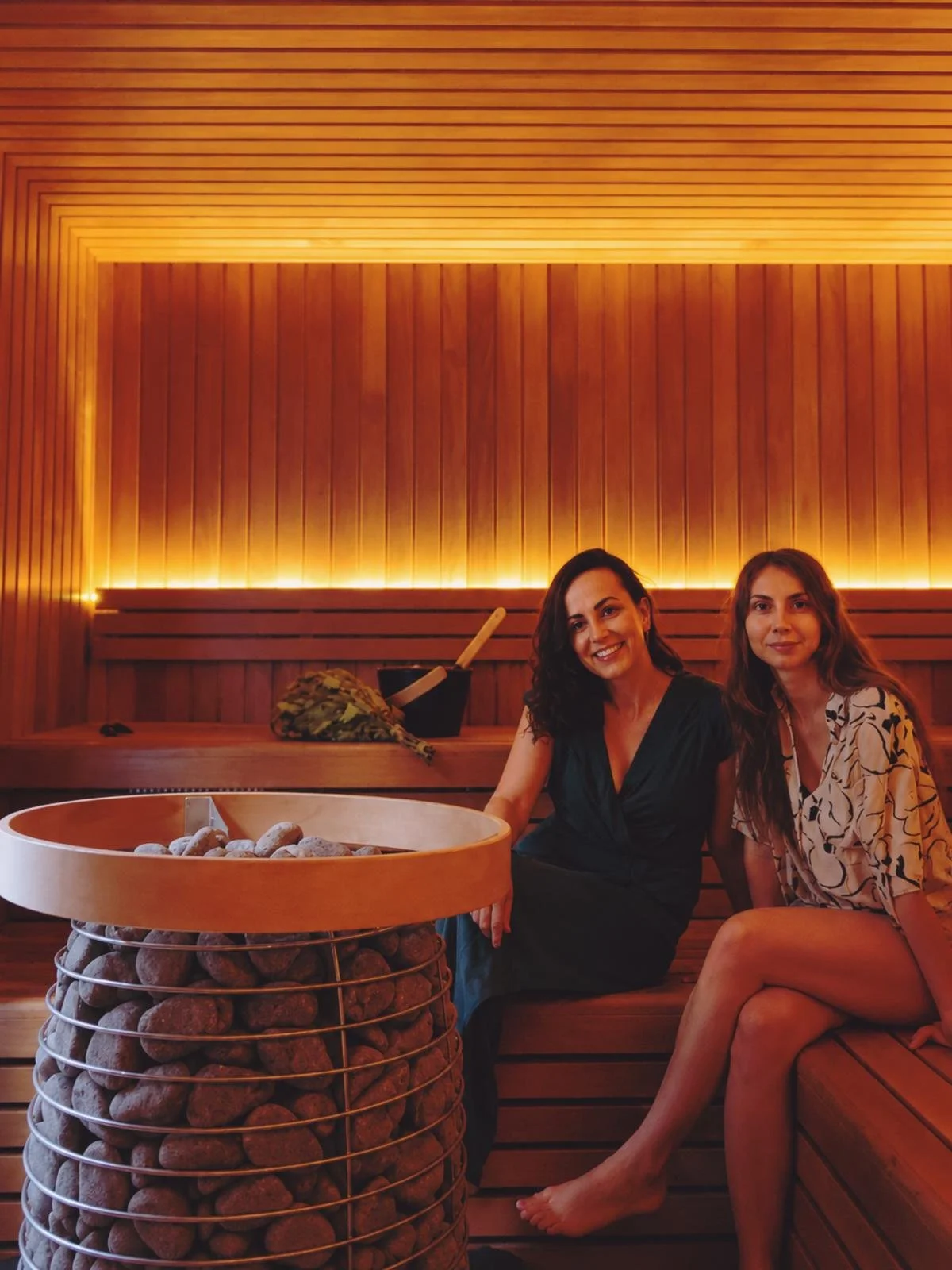 Two women sitting in a wooden sauna room, smiling, with a bucket of stones and sauna accessories nearby. Warm lighting enhances the relaxing atmosphere.