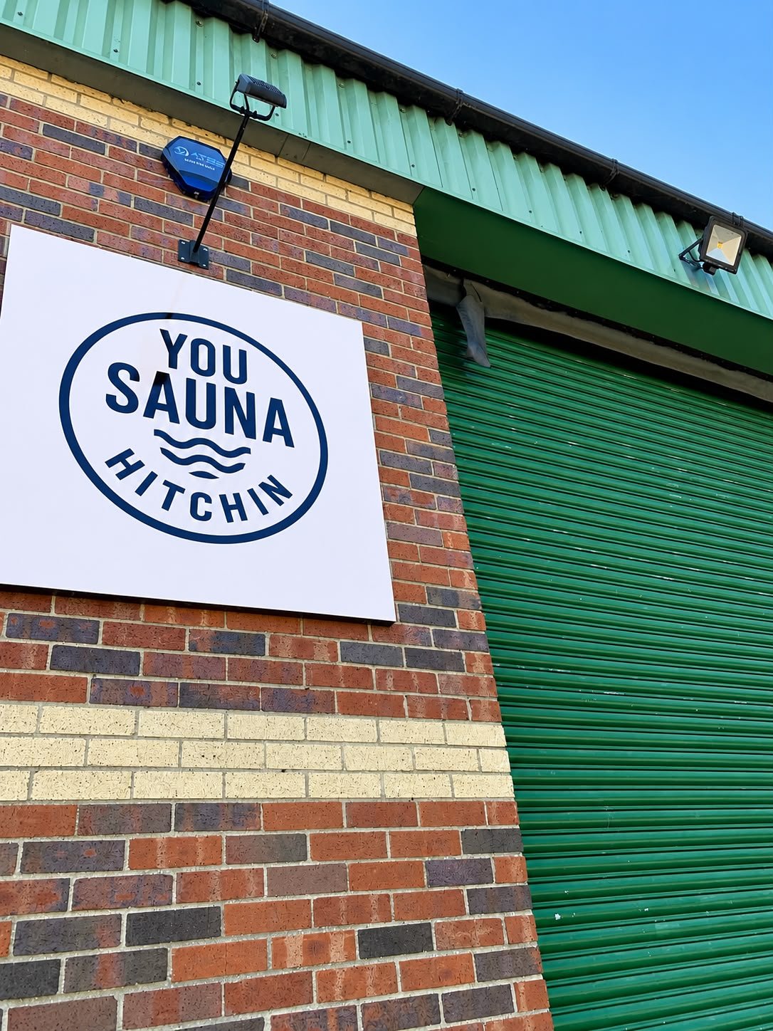 Exterior of a brick building with a white sign reading 'You Sauna Hitchin.' The building has a green metal roof and a green roll-up door.