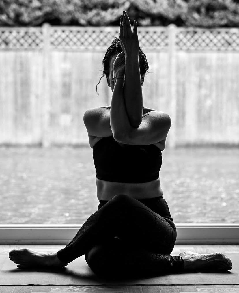 A woman practicing yoga indoors, sitting cross-legged with one arm raised and her hand covering her face, in front of a large window with an outdoor view.