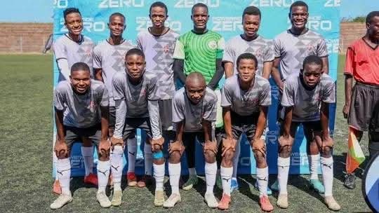 Group of ten young male soccer players in gray jerseys and black shorts, standing on a soccer field with a blue background and a referee in red to the right.