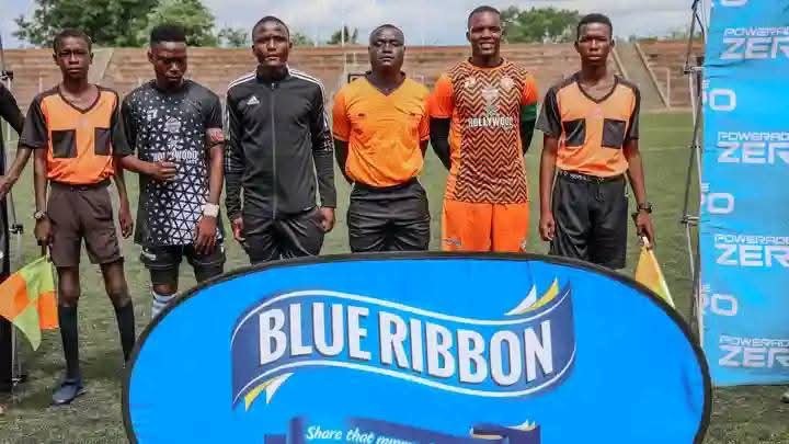 Group of six men standing on a football field behind a blue banner with 'Blue Ribbon' logo, some wearing black and orange sports uniforms, others in referee attire, with goalposts and stadium seating in background.