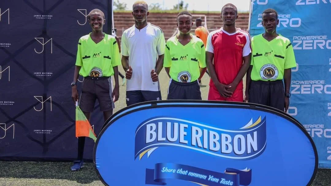 Four young soccer players in green jerseys and two adults in sports attire standing on a soccer field during a game day event with blue banners and a 'Blue Ribbon' sign in the foreground.