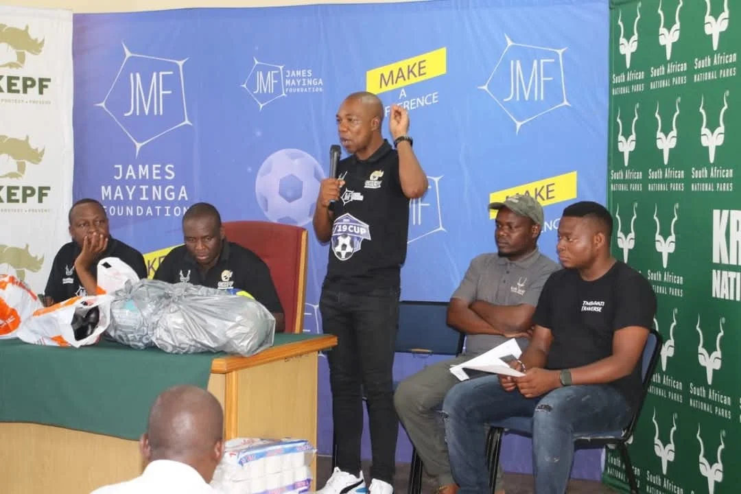 A man speaking into a microphone during a press conference, seated at a table with four other people, in front of a blue and green backdrop with logos for the James Mayinga Foundation, GKEPF and South African National Parks.