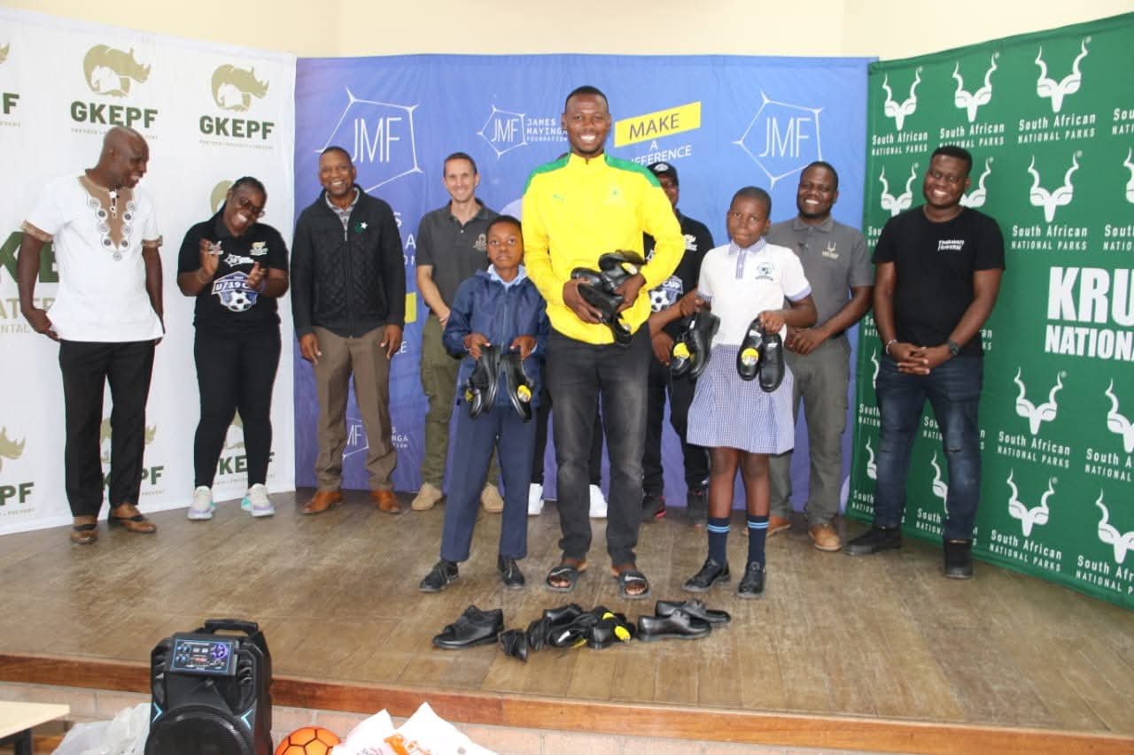 Group of people standing on a stage, some holding donated shoes, with banners of South African National Parks, GKEPF and James Mayinga Foundation in the background.