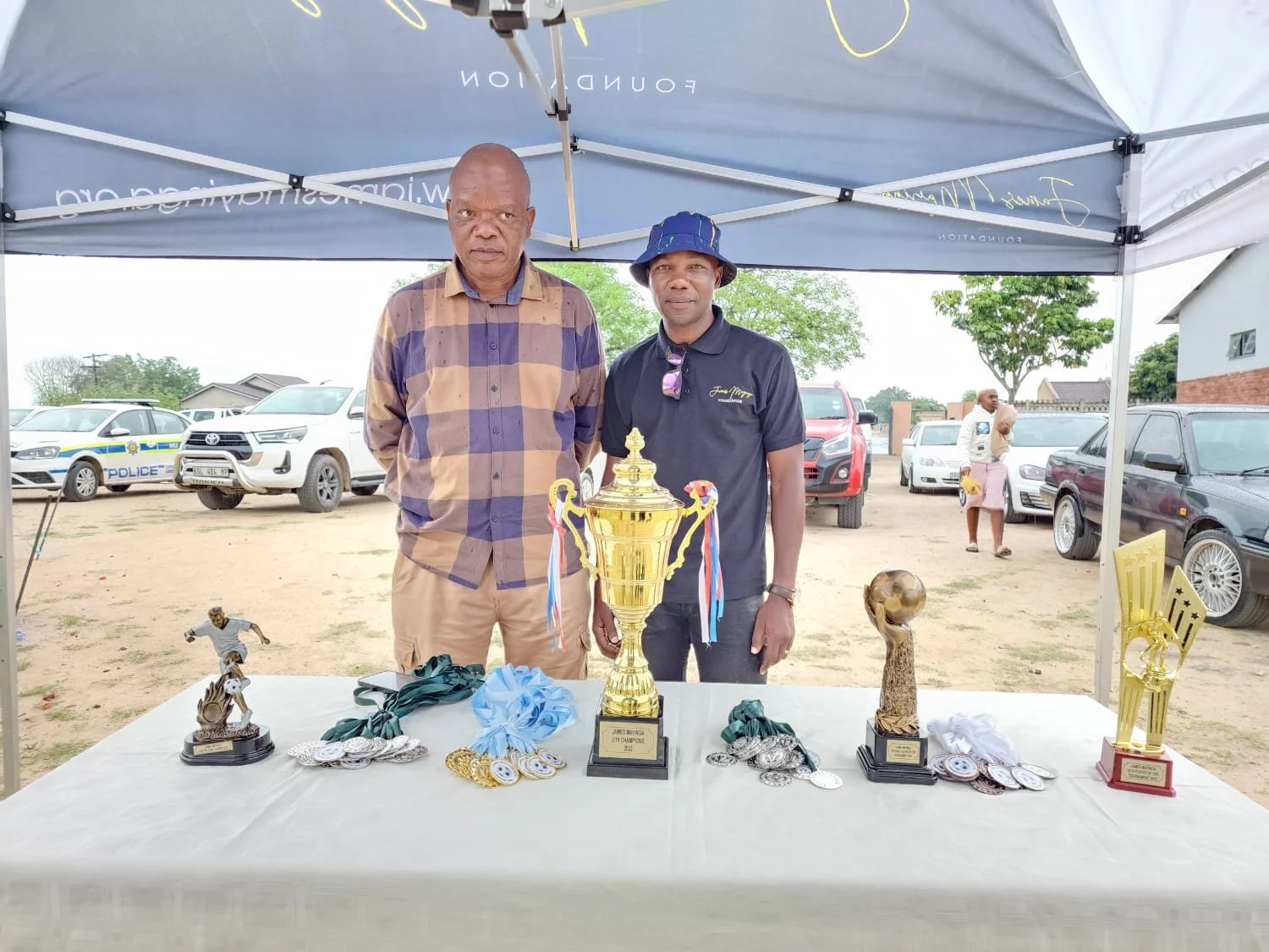 Two men standing behind a table filled with trophies and medals, under a canopy, with several parked cars and a few trees in the background.