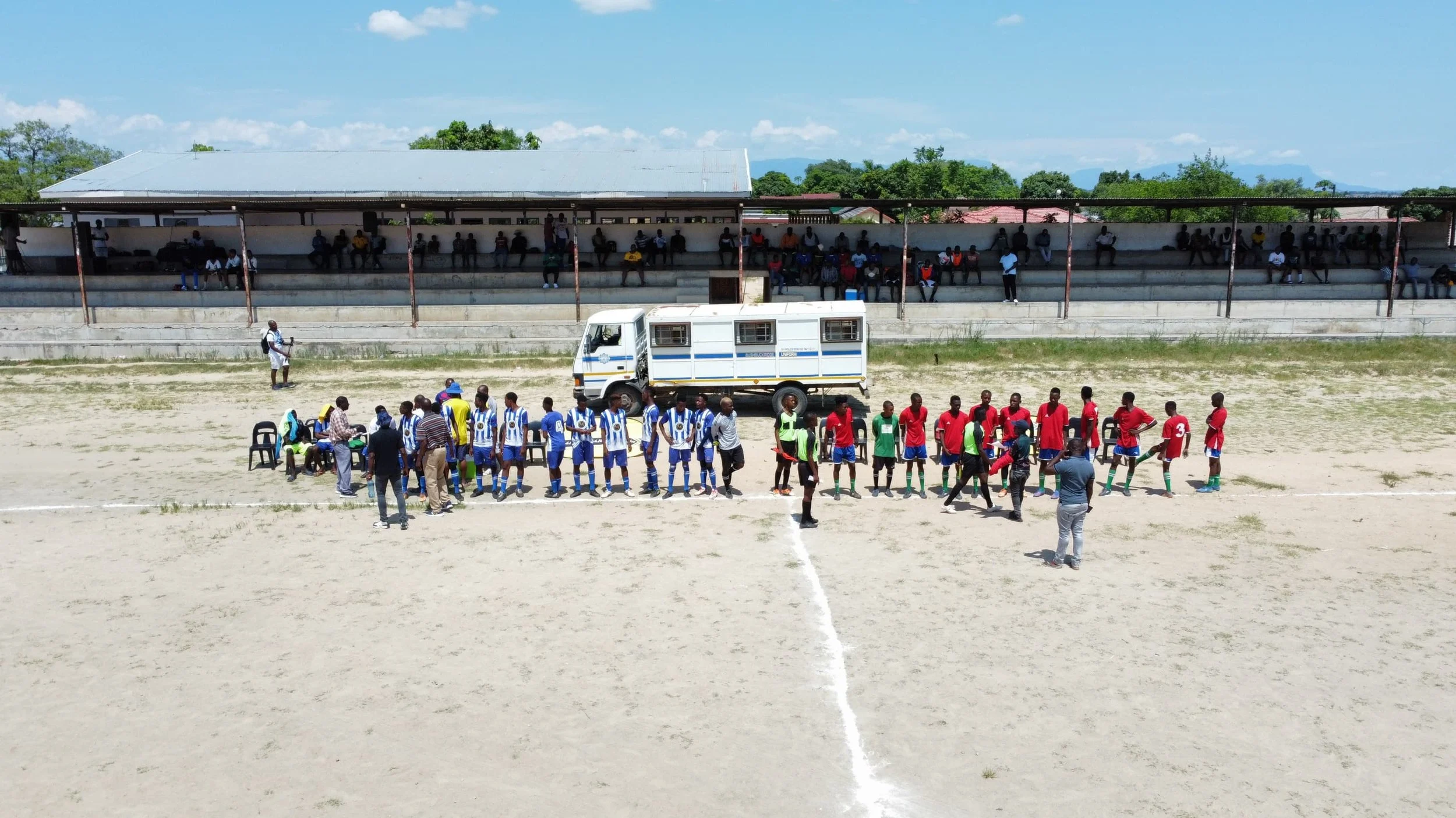Soccer match on a dirt field with players, referees, and officials, spectators seated in the stands, and a vehicle parked on the sidelines under a sunny sky.