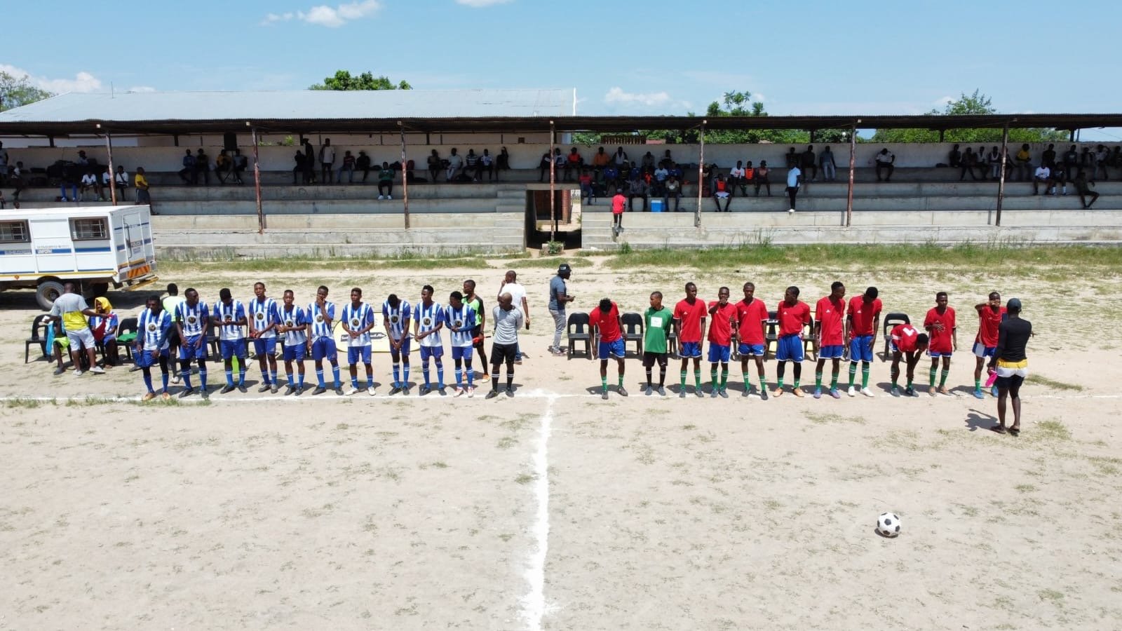 Two soccer teams in blue and red uniforms standing in a line on a dirt field, with spectators seated in a covered grandstand in the background.