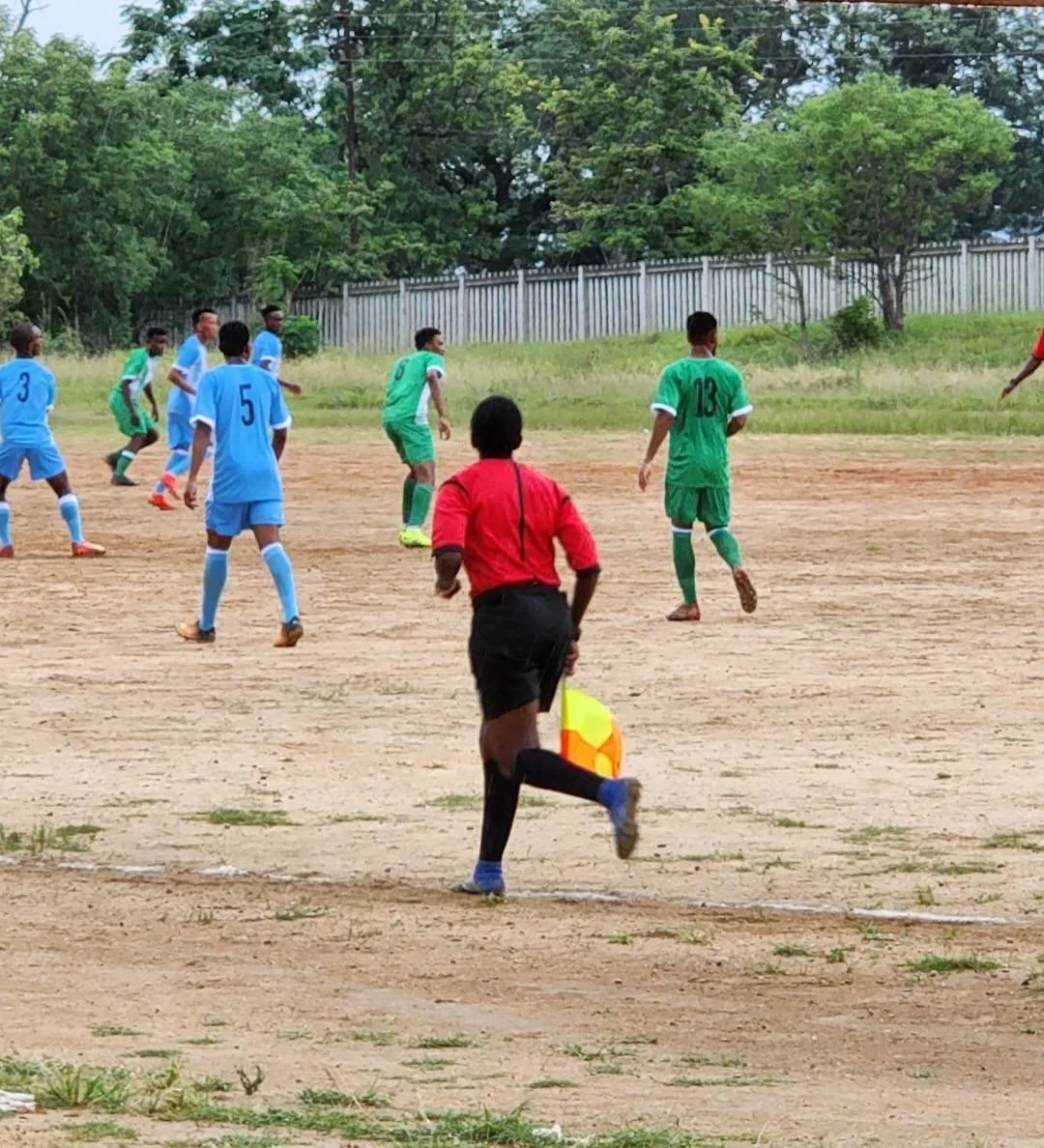 A soccer match with players in green and blue uniforms on a dirt field, with a referee running nearby holding a yellow and orange flag, and trees and a fence in the background.
