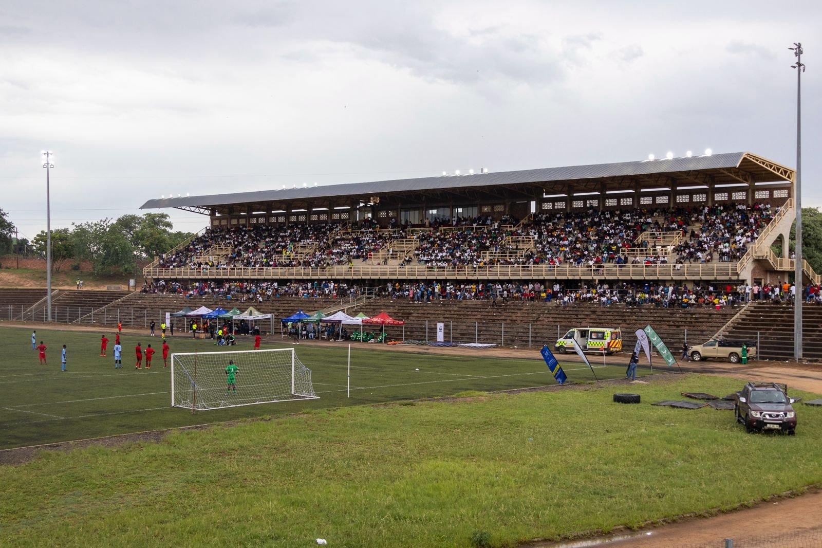 A football stadium filled with spectators watching a match, with players on the field and tents set up along the sideline.