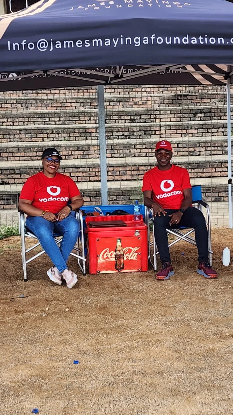 Two smiling people sitting under a canopy with a Coca-Cola cooler in front. They are wearing red Vodacom shirts and caps, with chairs and water bottles nearby.
