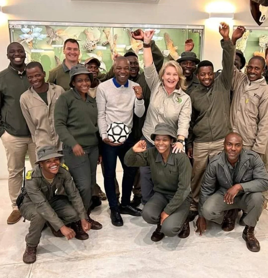 Group of smiling people, some in ranger uniforms, celebrating indoors with a woman holding a soccer ball, raising fists, and surrounded by others.