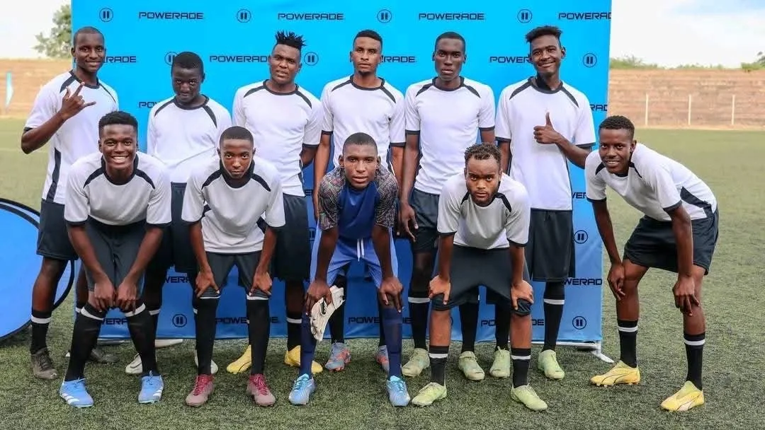 Group of young male soccer players in white and black uniforms posing on a soccer field, with a blue Powerade branded backdrop behind them.