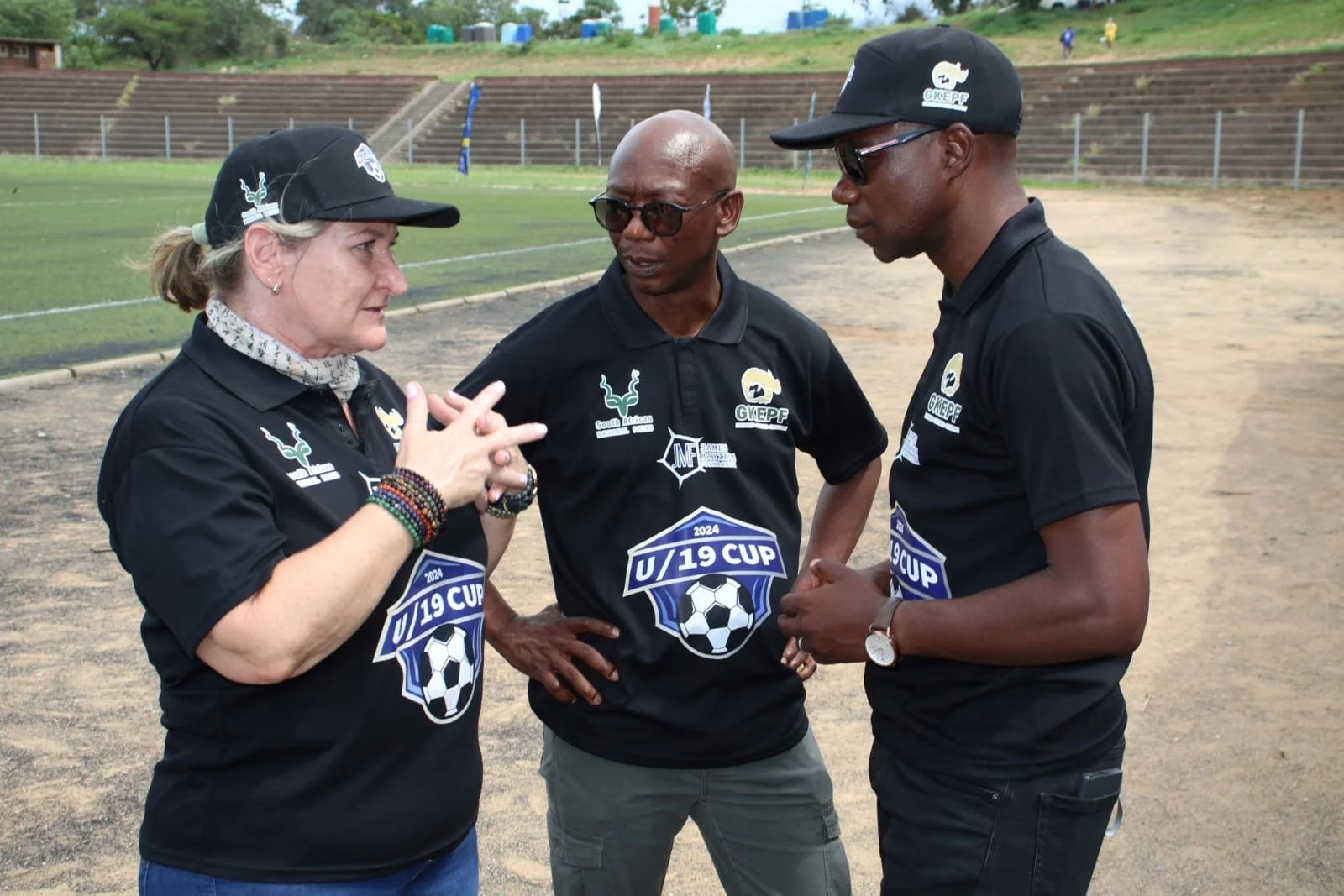 Three people wearing black shirts with event logos, talking on a soccer field with bleachers and trees in the background.