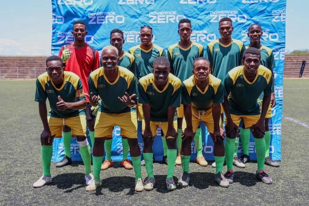 Eleven male soccer players pose on a field in front of a blue backdrop with the words "Powerade Zero" printed on it. They are wearing green and yellow uniforms, with one player in a red goalkeeper jersey. The players are smiling and standing in two r