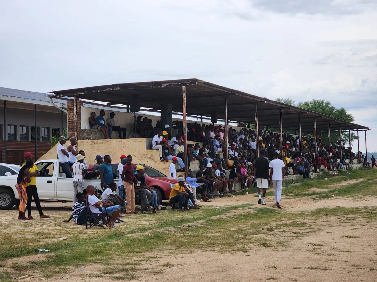 Crowd of people gathered in an outdoor stadium with some seated under a large shelter and others sitting on cars and chairs outside on a dirt and grass field.