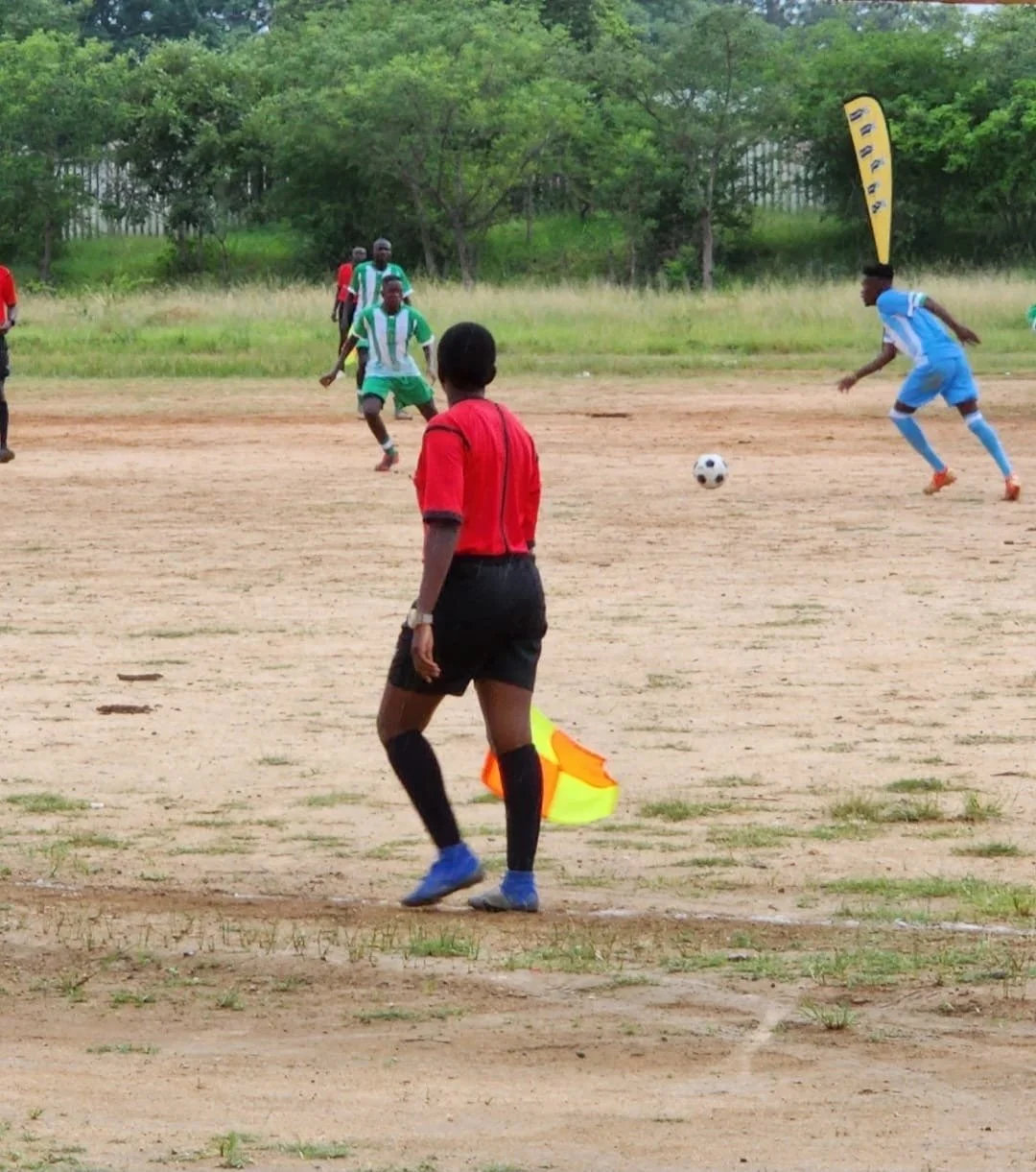 A soccer match on a dirt field with players in colorful uniforms and a referee in a red shirt and black shorts, near a line of trees.