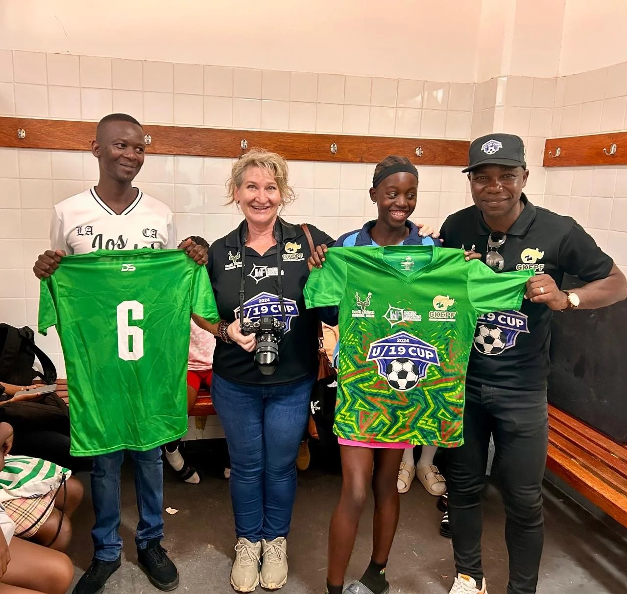 Four people standing in a locker room with two women and two men, holding green soccer jerseys with the text 'U/19 Cup 2024' and a soccer ball graphic.