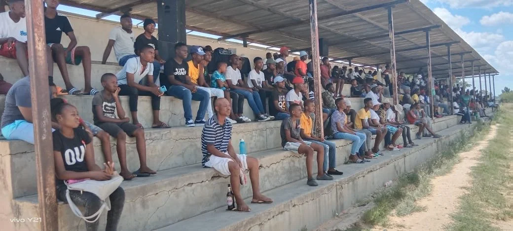 A large group of children and some adults sitting on concrete bleachers under a roof, watching an event outdoors on a sunny day.