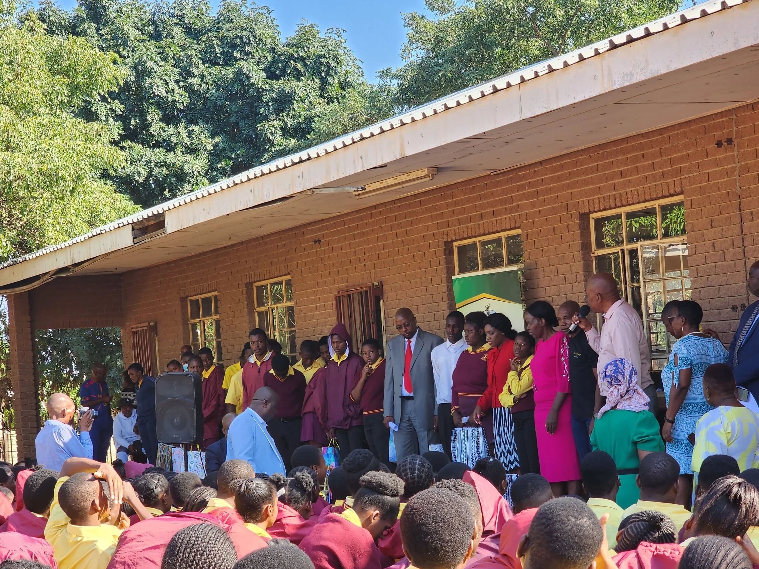 A group of students and adults standing on a stage outside a brick building, with a large crowd seated in front. There is a man speaking into a microphone, and the scene is set in a sunny outdoor area with trees in the background.