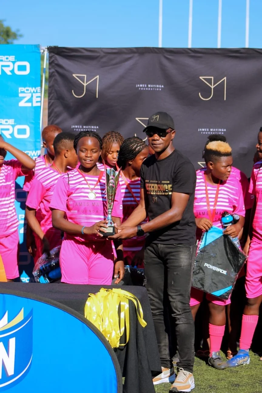 A man presents a trophy to a young girl in a pink sports uniform during an outdoor sports award ceremony, with other girls in pink uniforms and a backdrop featuring the James Mavinga Foundation logo.