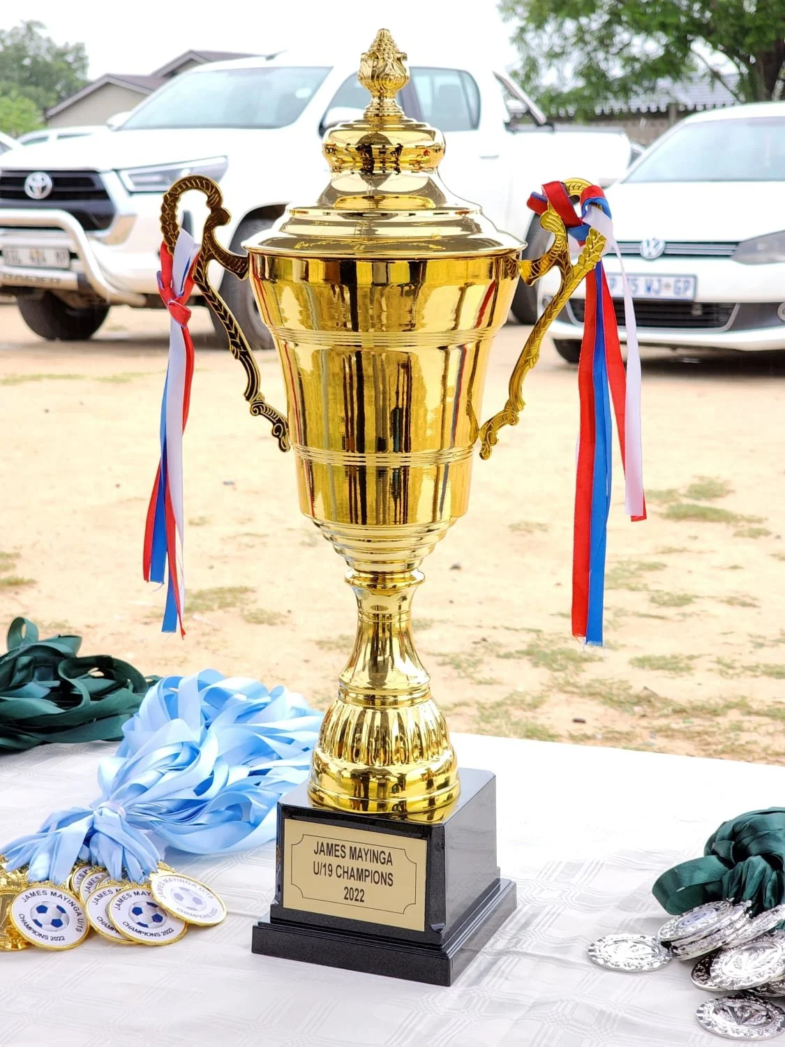 Gold championship trophy on a table with medals, ribbons, and cars in the background, awarded for James Mayinga U19 Champions 2022.