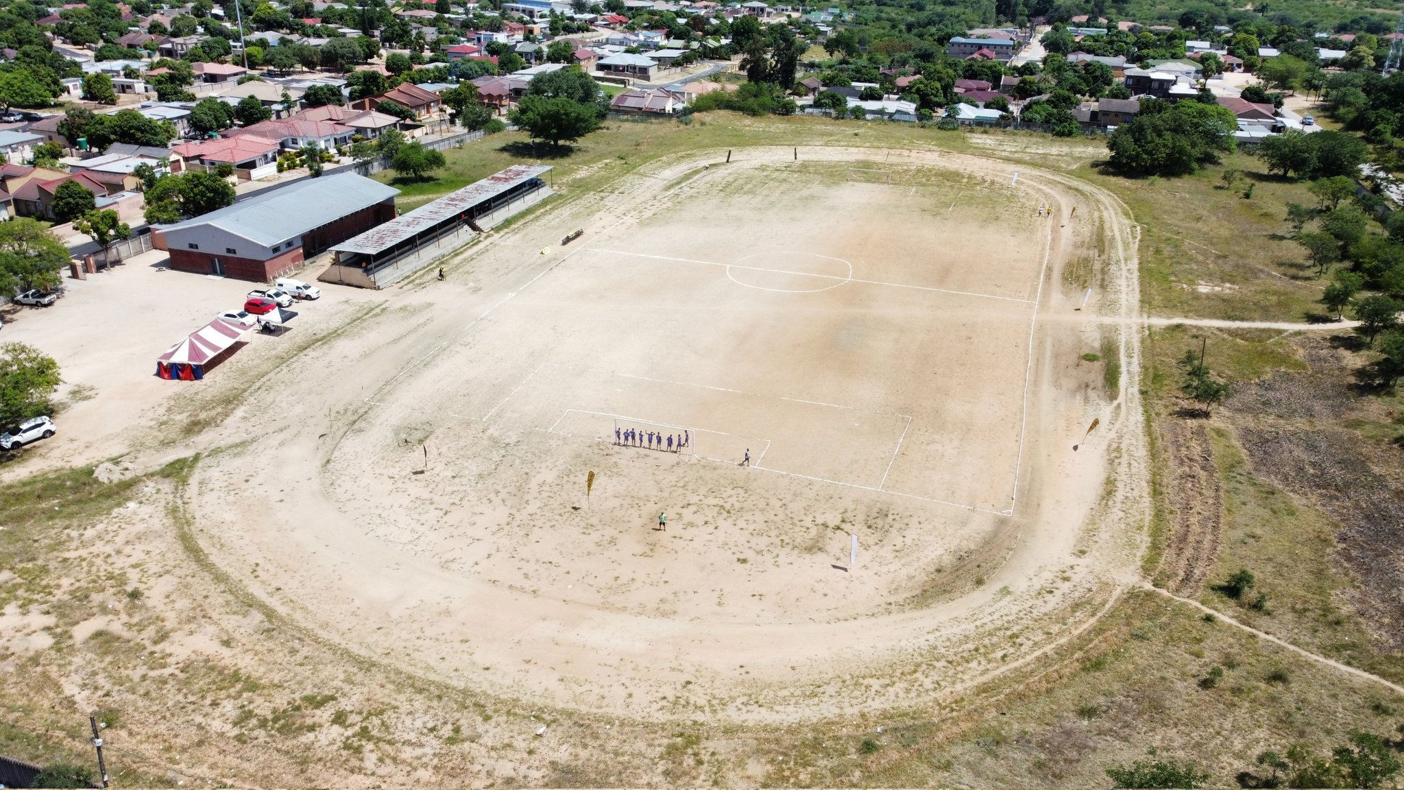 An aerial view of a dirt football field. The field has a small group of people standing near the goal and some others scattered around. There are tents and parked cars on one side of the field.