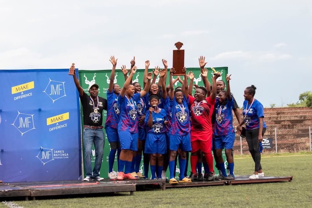 A group of young female players celebrating on a stage field with medals, holding a trophy, and smiling for the photo.