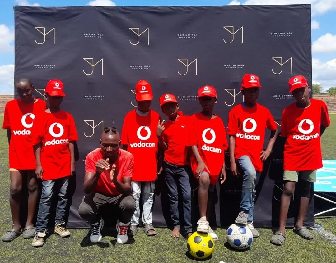 Group of young boys and an adult man posing on a soccer field in front of a black backdrop with logos. They are wearing red jerseys and caps with Vodafone and Dacom branding, and some have soccer balls at their feet.
