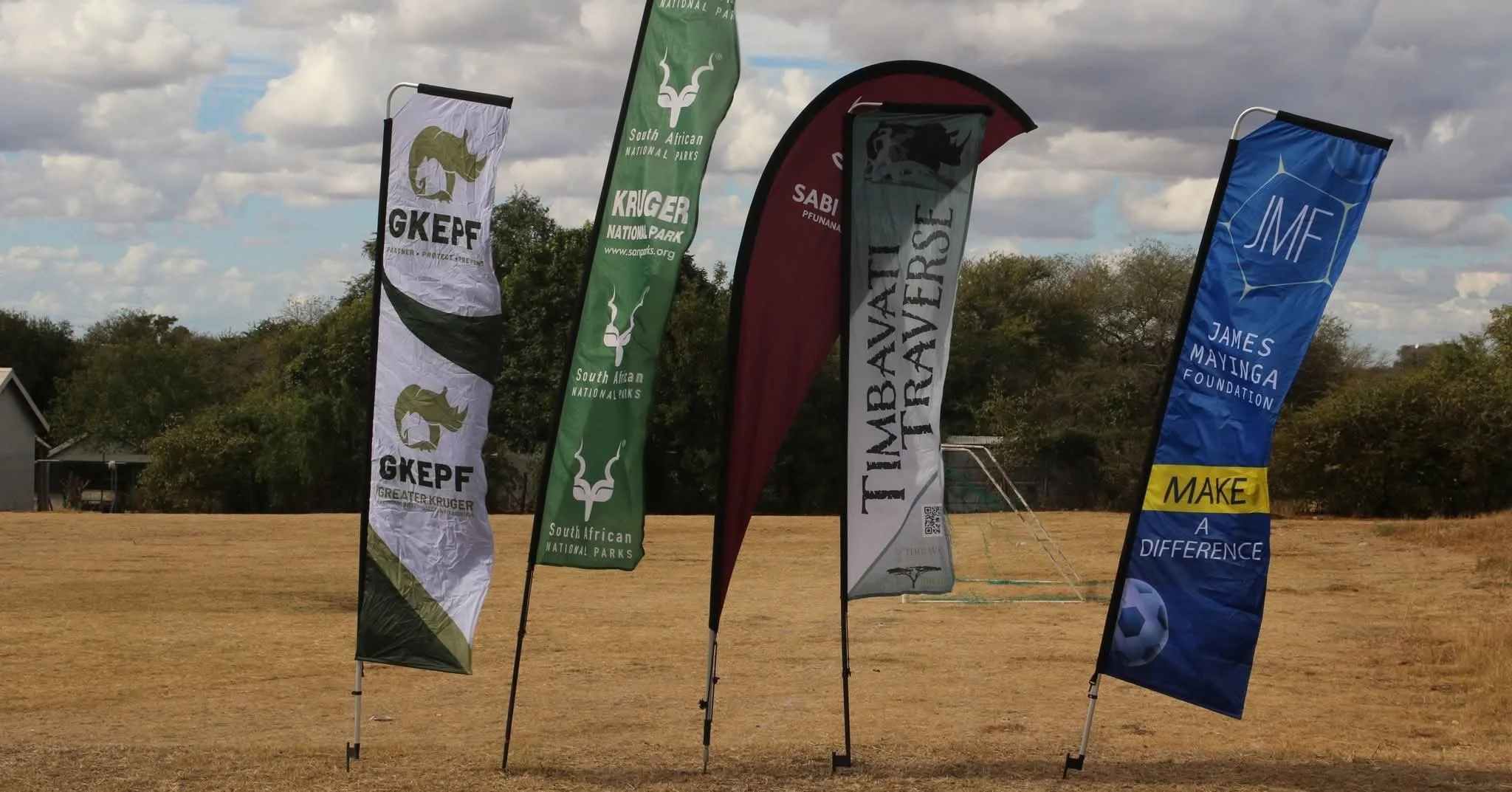 Five colorful flags on a grassy field with trees and a cloudy sky in the background. The flags display logos and text for various organizations and foundations, including GKEPF, South African National Parks, Timbavati Transfrontiers, and James Mayinga Foundation.