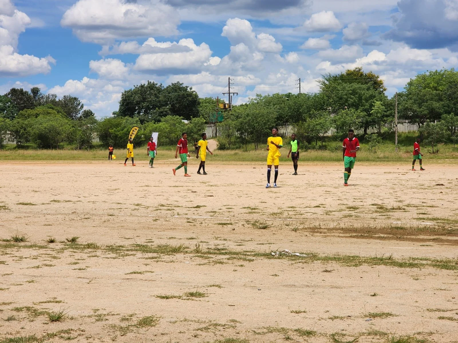 Soccer game on a dirt field with players wearing red, yellow, and green uniforms under a partly cloudy sky.