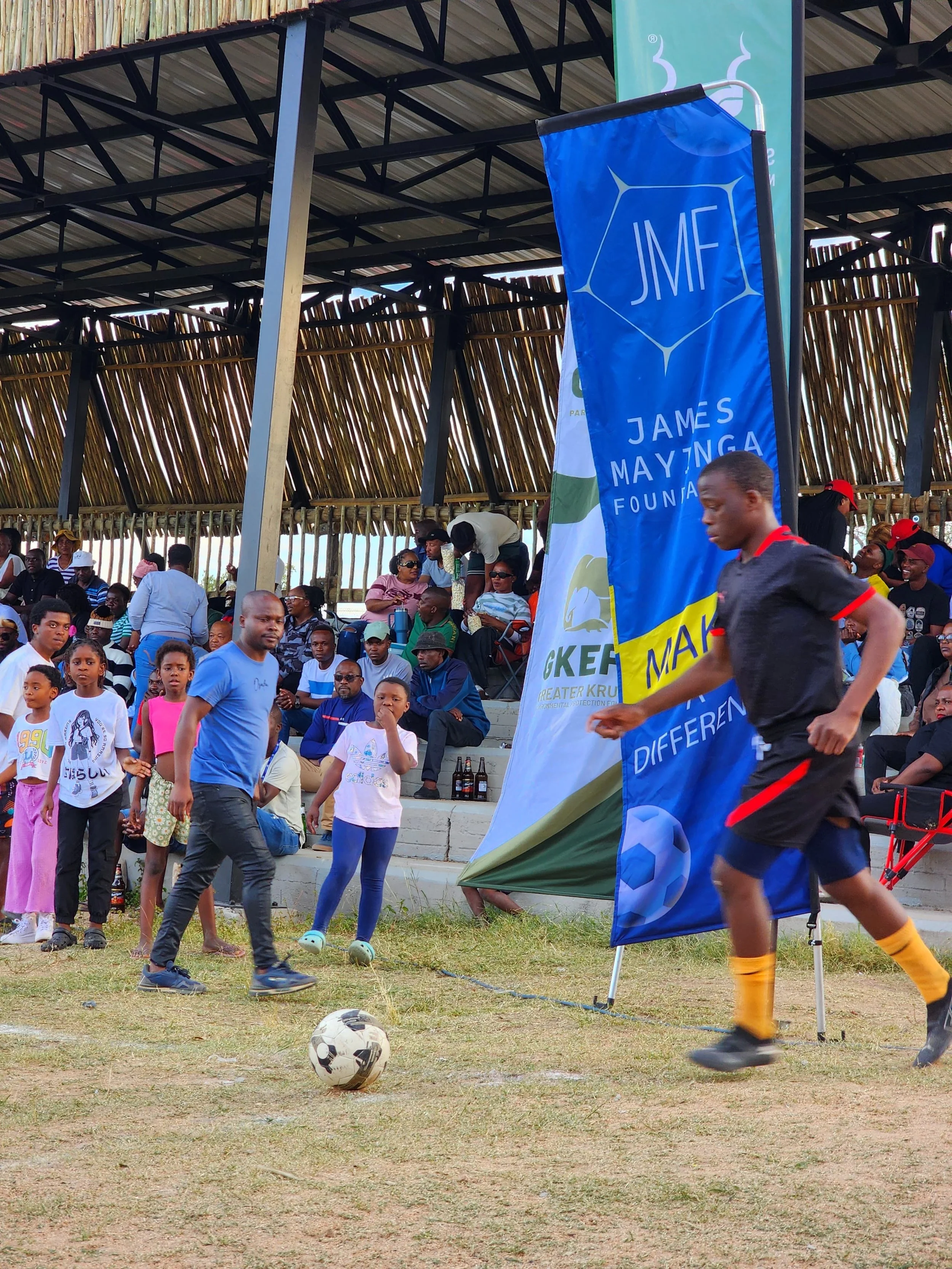 People watching a soccer game at an outdoor stadium, with children and adults standing near the sidelines, and a player running past with a soccer ball, during a daytime event.