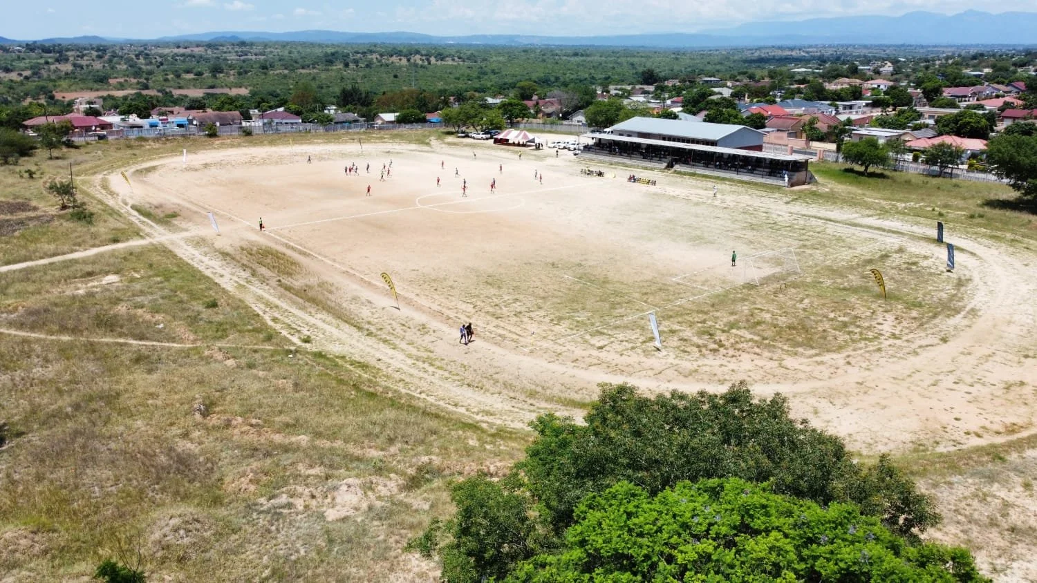 An aerial view of a soccer field with kids playing, surrounded by a dirt running track, with bleachers, small building, and residential houses in the background.