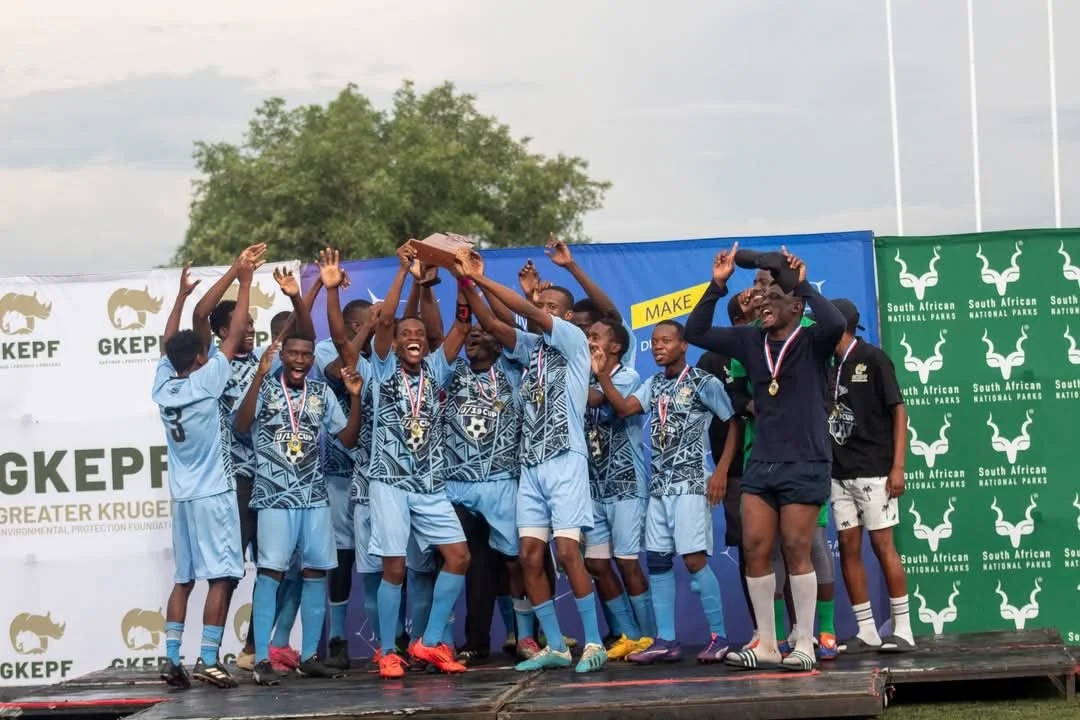 A group of young soccer players celebrating on a podium, holding a trophy, wearing medals, and smiling.