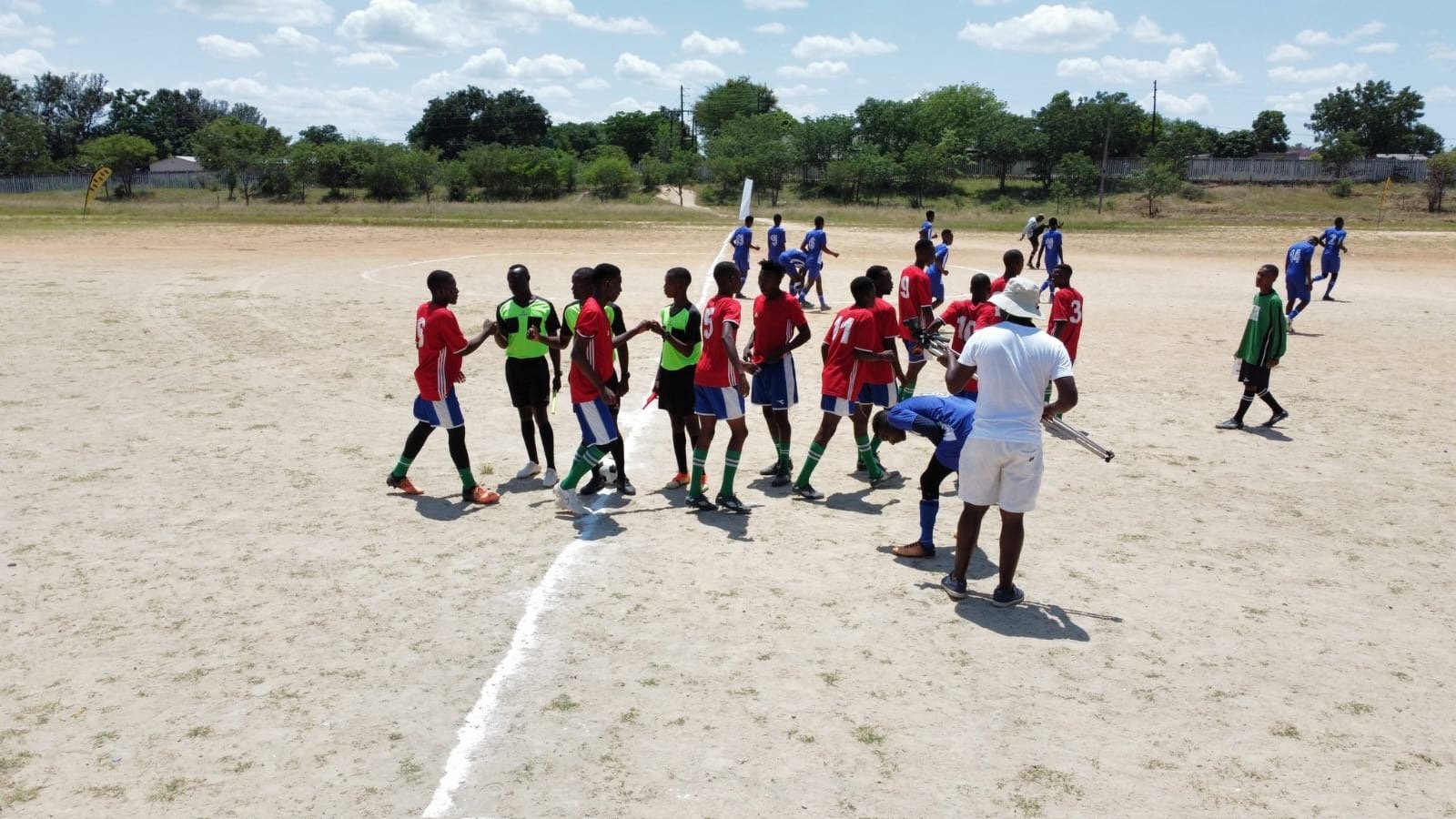 Young soccer players in red and blue uniforms exchanging handshakes on a dusty field with some players in blue uniforms in the background.