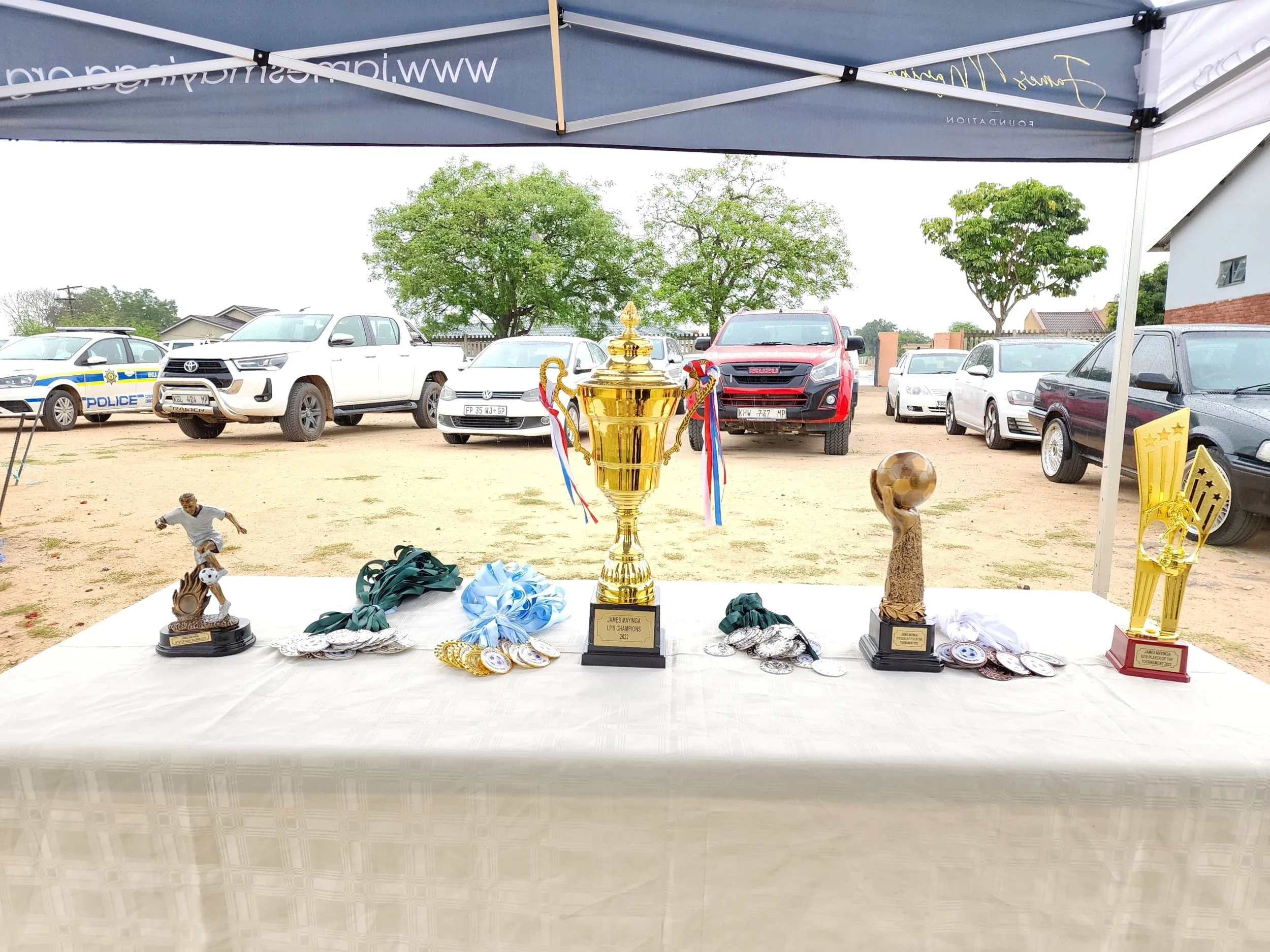 Table with multiple medals and trophies in an open outdoor space, with parked cars and trees in the background, under a canopy.