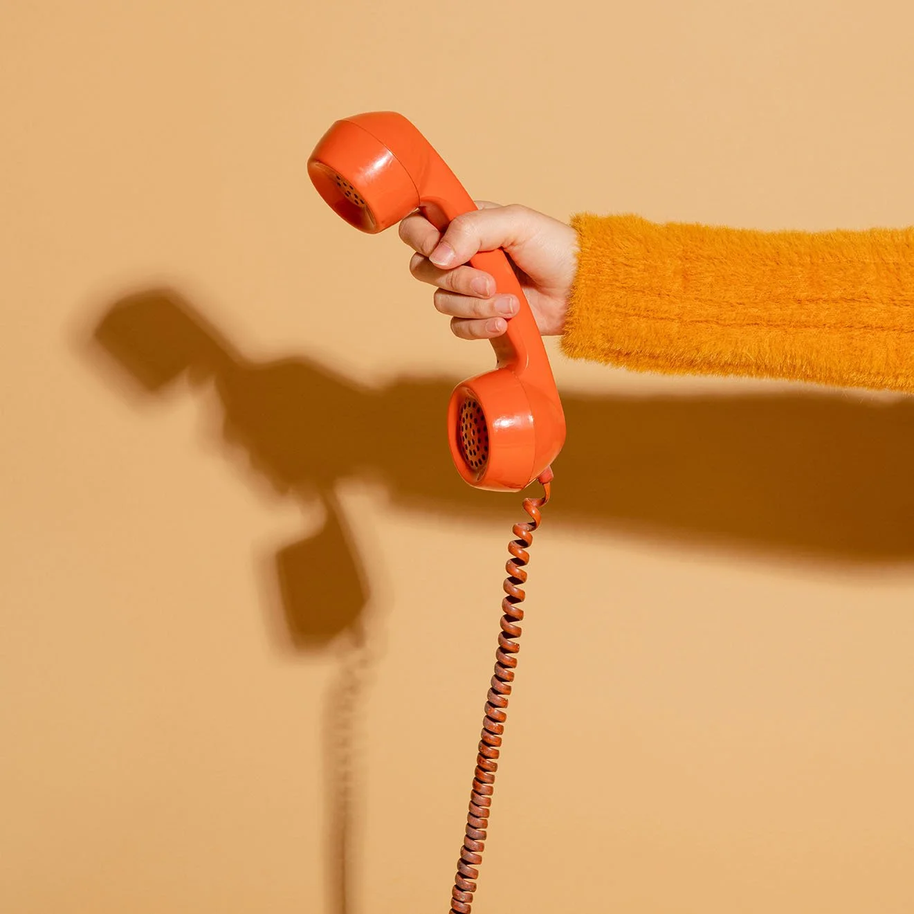 A person wearing an orange fuzzy sweater holds a vintage orange corded telephone, casting a shadow on a beige wall.