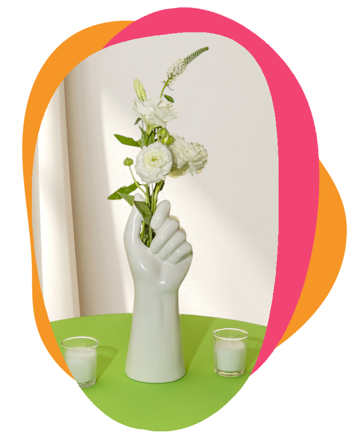 Decorative arrangement of white flowers in a ceramic hand-shaped vase, flanked by two small white candles on a green table against a plain white wall.