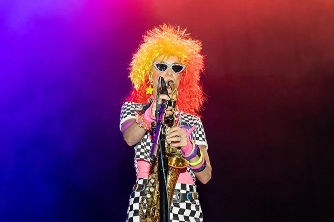 Person with curly yellow and orange hair wearing sunglasses, holding a microphone and a saxophone, dressed in a colorful checkered outfit and jewelry, on stage with vibrant purple and red lighting.