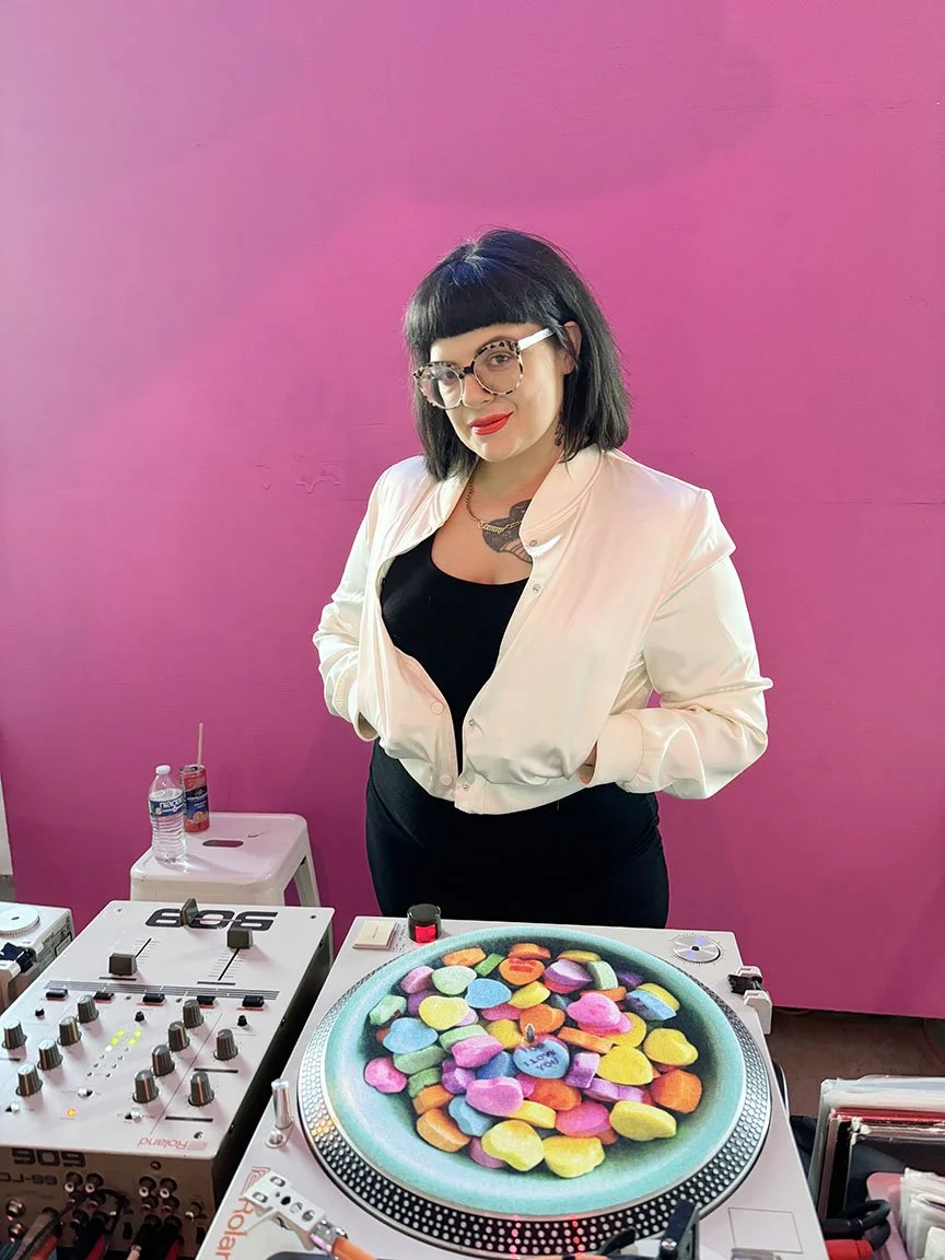 A woman with black hair, glasses, and red lipstick stands behind a DJ turntable with colorful stones or candies arranged in a circular pattern on top of it. She is wearing a white jacket over a black top, standing against a pink background.
