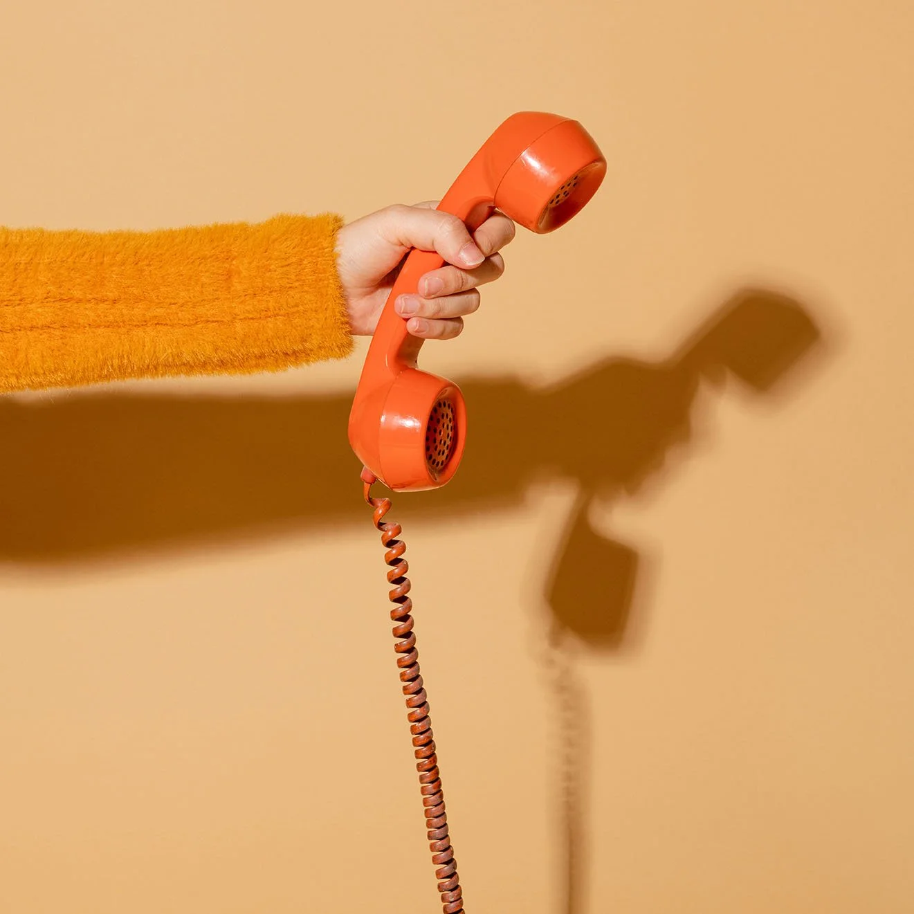 A person with a fuzzy orange sleeve holding an orange vintage corded telephone against a beige background, casting a shadow.