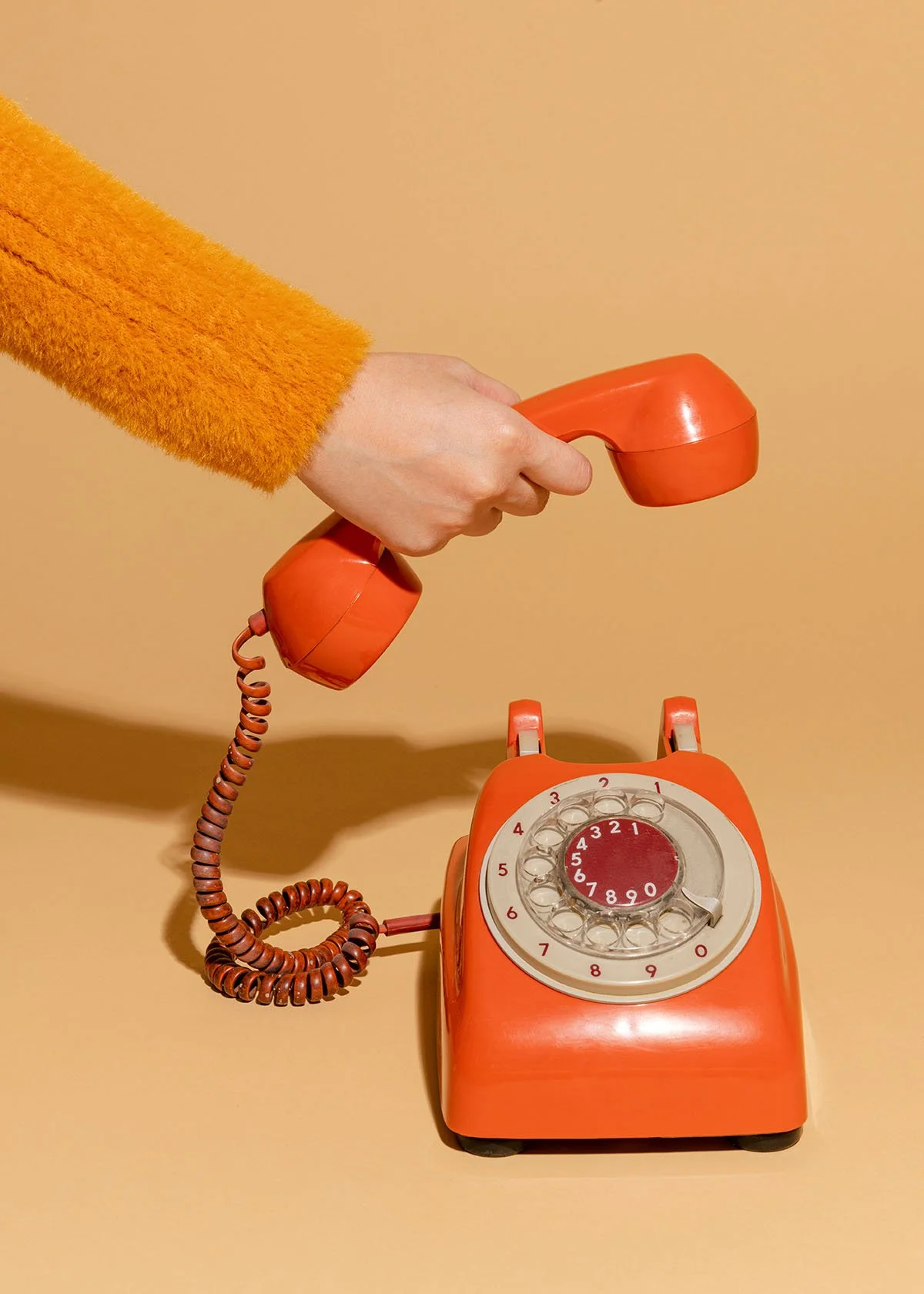 A person wearing an orange fuzzy sweater holding a vintage orange rotary telephone with the receiver in their hand, positioned above an orange rotary dial phone on a beige background.