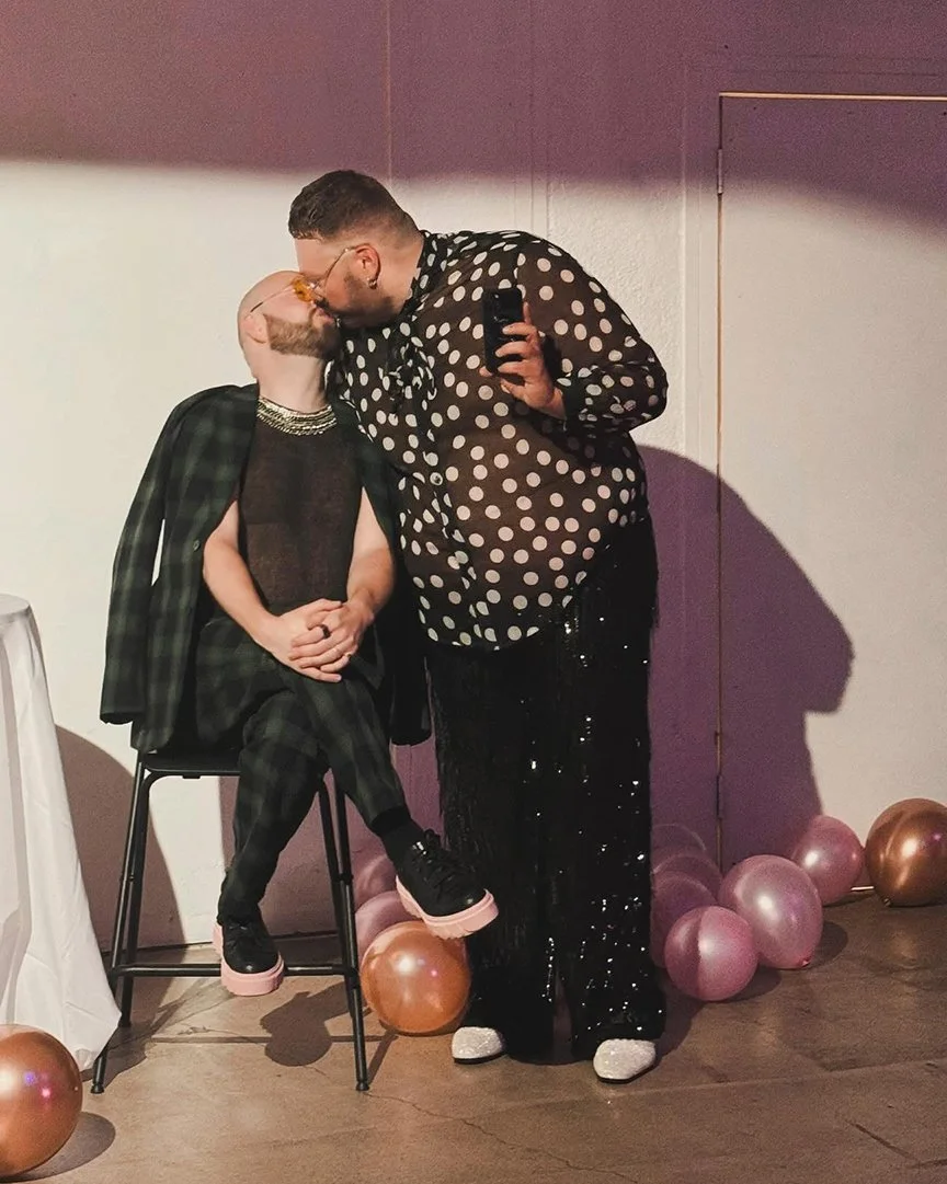 Two people sharing a kiss, one sitting on a stool wearing a black plaid jacket and pants, and the other standing in a polka dot shirt and shiny pants. Balloons are on the floor around them, and the setting appears to be a party or celebration.