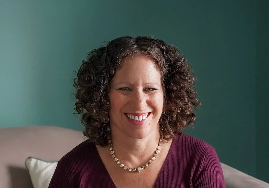 A smiling woman with curly brown hair, wearing a purple top and a pearl necklace, sitting on a beige chair with a white pillow, against a teal wall.