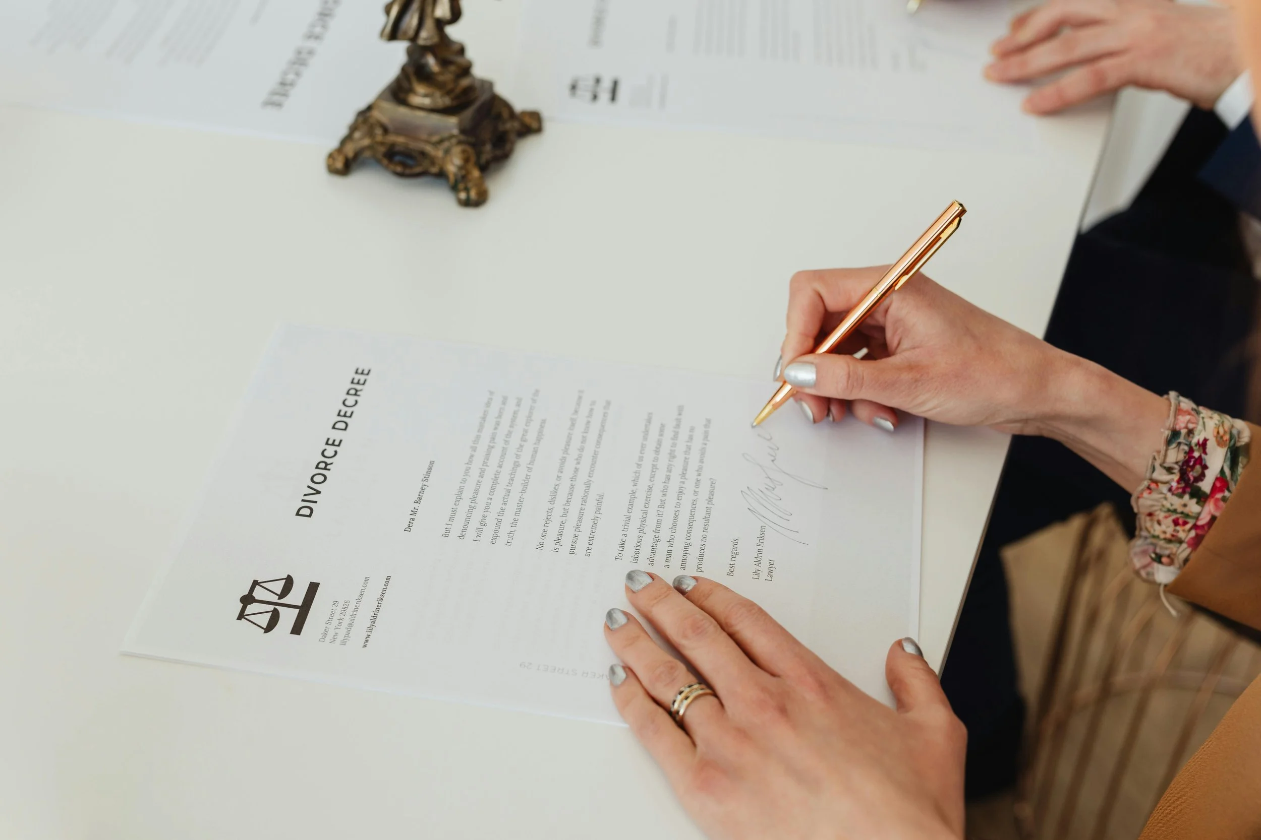 A person signing a divorce document with a gold pen, seated next to another person at a white table with a decorative brass sculpture and other papers.