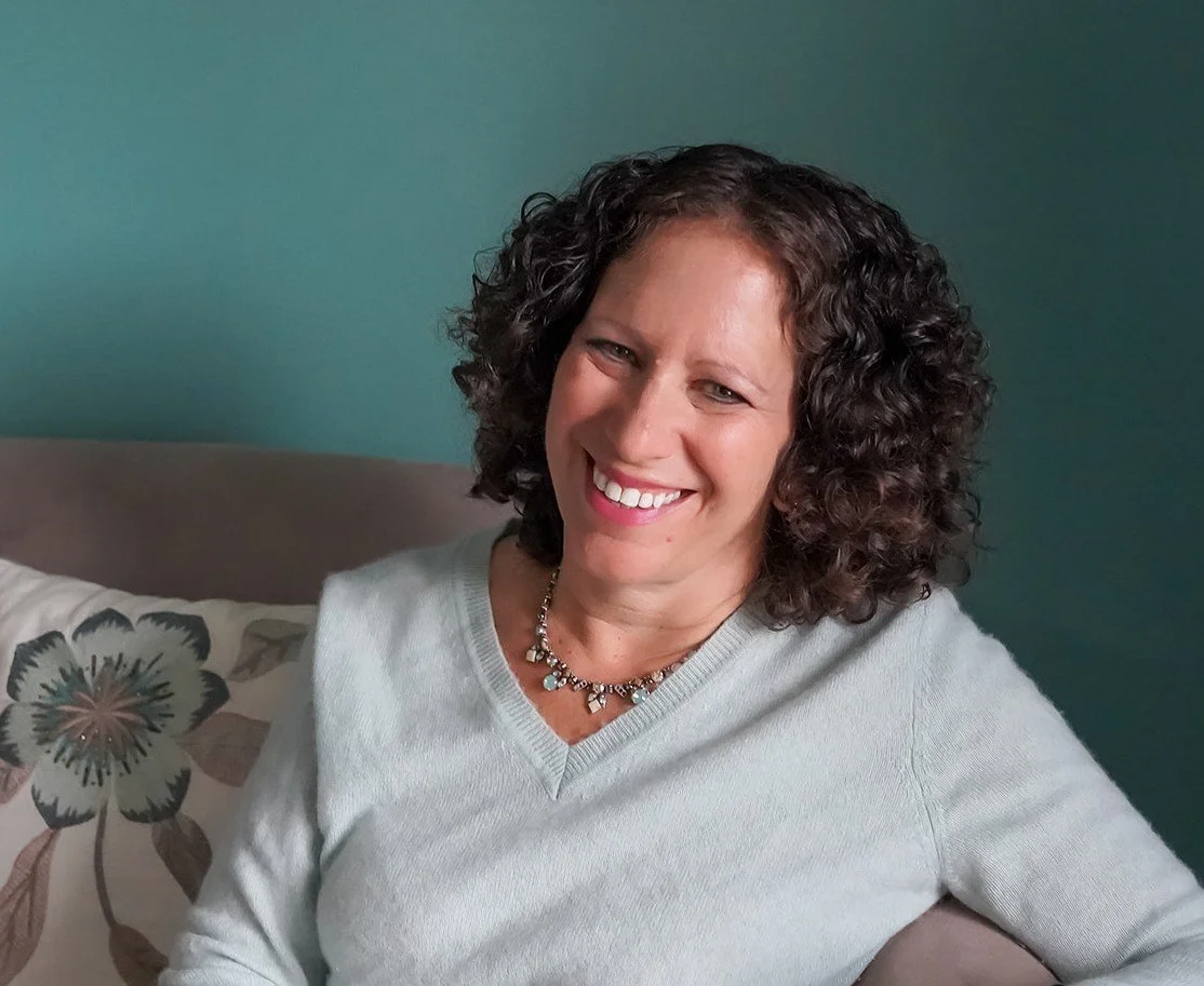 A woman with curly dark hair smiling, sitting on a couch with a floral pillow, wearing a light-colored sweater and a beaded necklace, against a green wall.