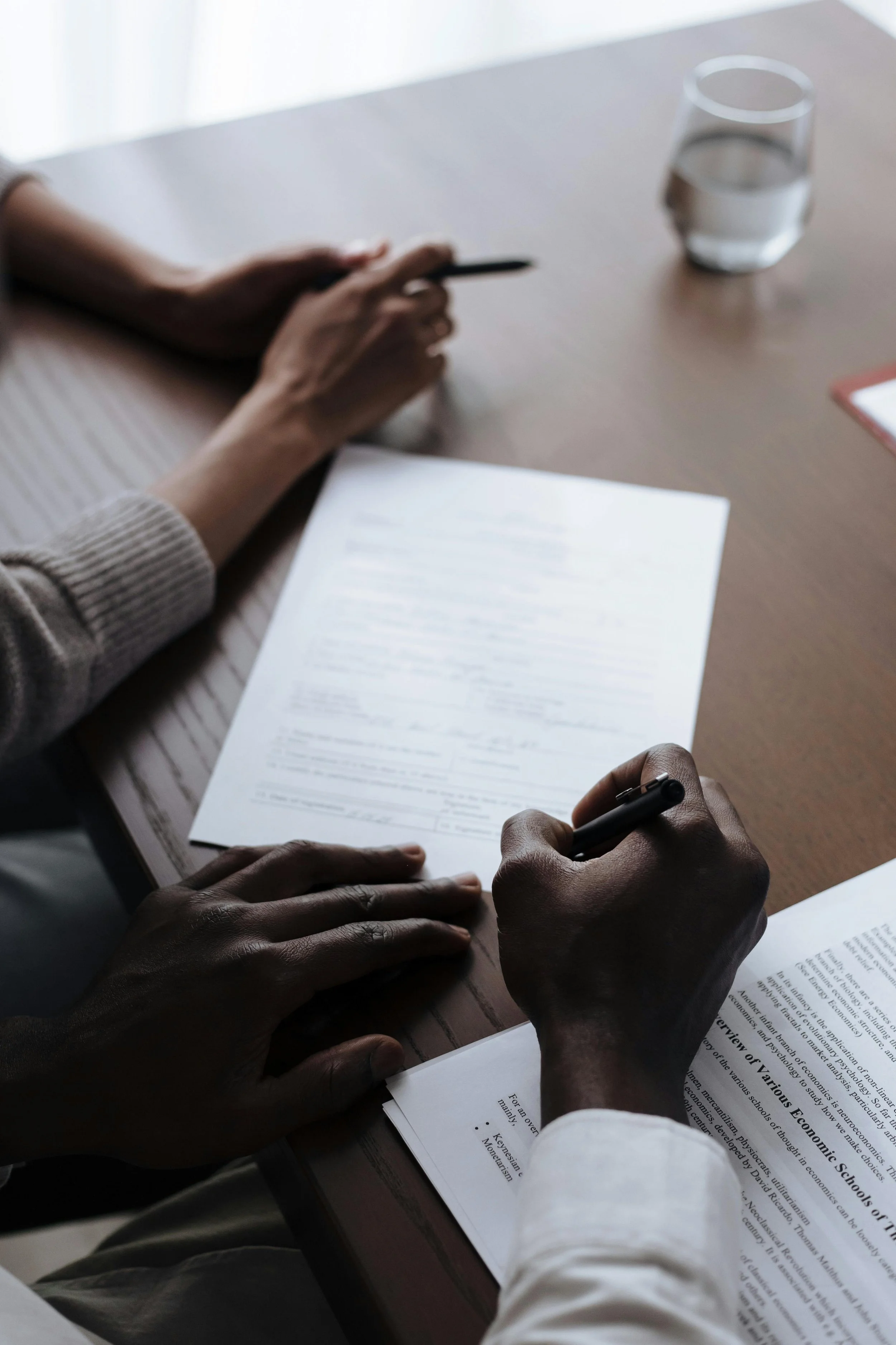 Close up view of two pairs of hands at a desk reviewing and signing divorce documents, with a glass of water nearby.